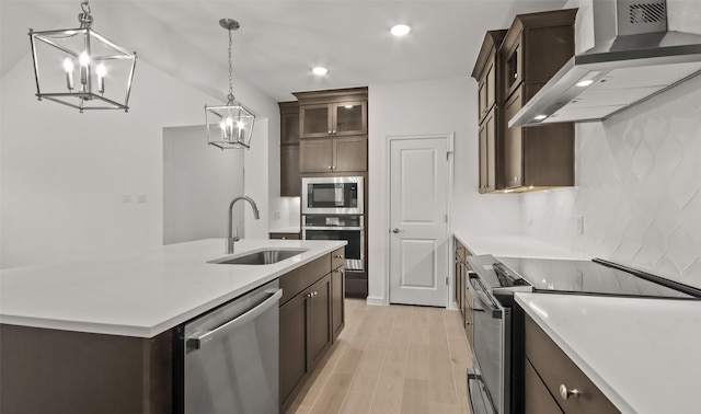 kitchen with sink, a center island with sink, hanging light fixtures, appliances with stainless steel finishes, and wall chimney range hood