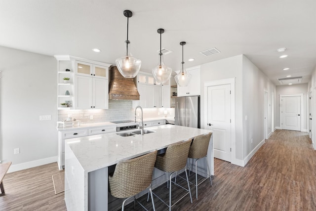 kitchen featuring premium range hood, a center island with sink, hanging light fixtures, dark hardwood / wood-style floors, and stainless steel fridge