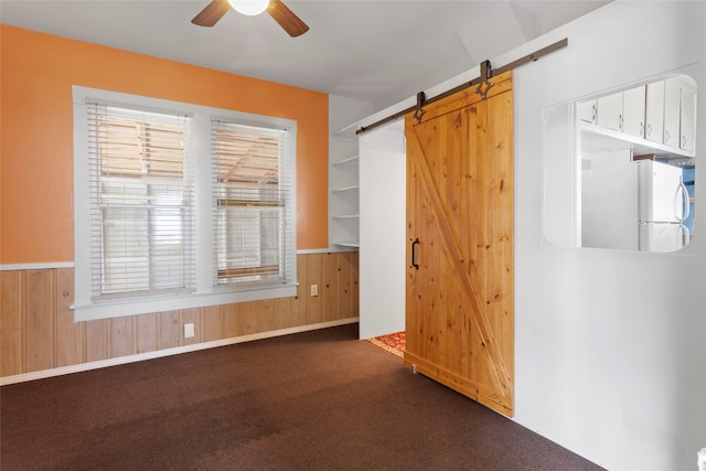 spare room featuring a barn door, wood walls, ceiling fan, and dark carpet