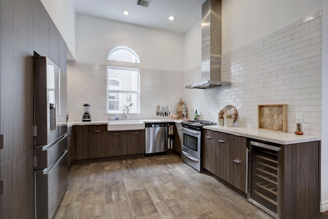 kitchen featuring decorative backsplash, appliances with stainless steel finishes, beverage cooler, sink, and wall chimney range hood