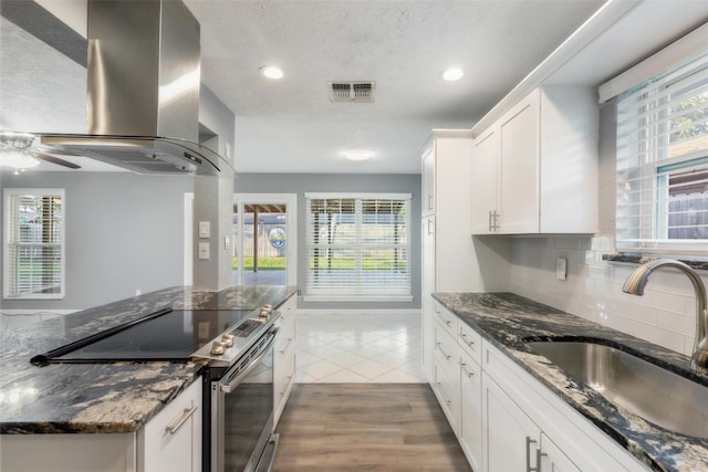 kitchen with sink, wall chimney exhaust hood, a healthy amount of sunlight, stainless steel range with electric stovetop, and white cabinets