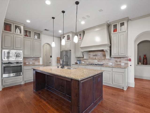 kitchen featuring sink, stainless steel appliances, premium range hood, backsplash, and a center island with sink