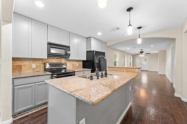 kitchen with ceiling fan, hanging light fixtures, light stone counters, an island with sink, and black appliances