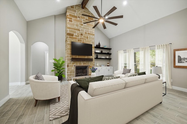 living room with beam ceiling, ceiling fan, a stone fireplace, high vaulted ceiling, and light hardwood / wood-style floors