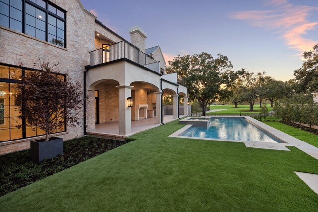 back house at dusk with a lawn, a patio area, a balcony, and a fireplace