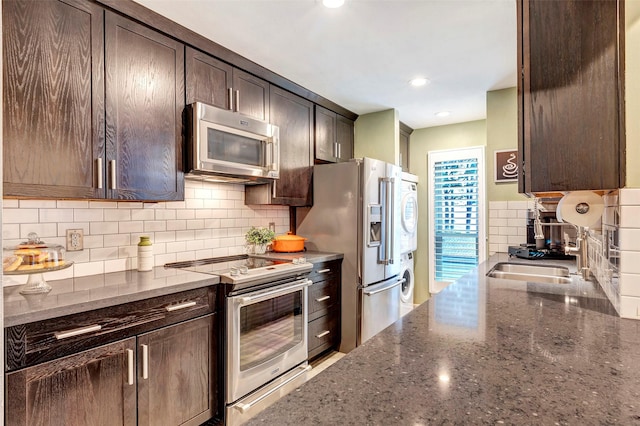 kitchen with dark brown cabinetry, sink, dark stone countertops, stacked washer and dryer, and appliances with stainless steel finishes