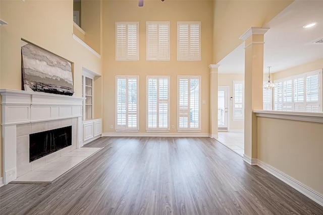 unfurnished living room with a towering ceiling, light wood-type flooring, ornate columns, a notable chandelier, and a tiled fireplace