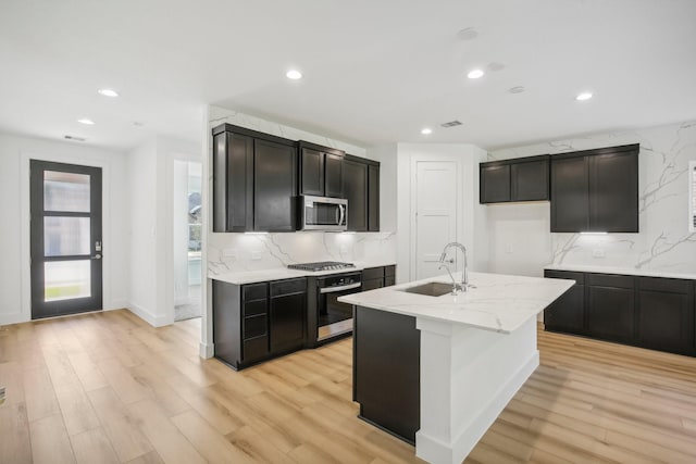 kitchen with sink, stainless steel appliances, an island with sink, decorative backsplash, and light wood-type flooring