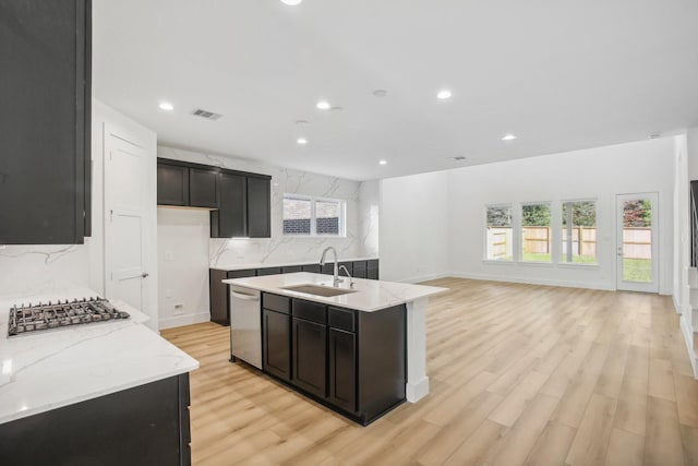 kitchen with light stone countertops, sink, stainless steel appliances, a kitchen island with sink, and light wood-type flooring