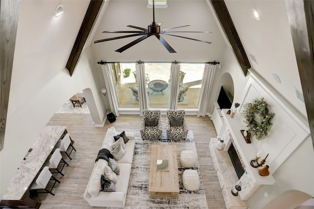 living room featuring high vaulted ceiling, a stone fireplace, light hardwood / wood-style flooring, ceiling fan, and beam ceiling