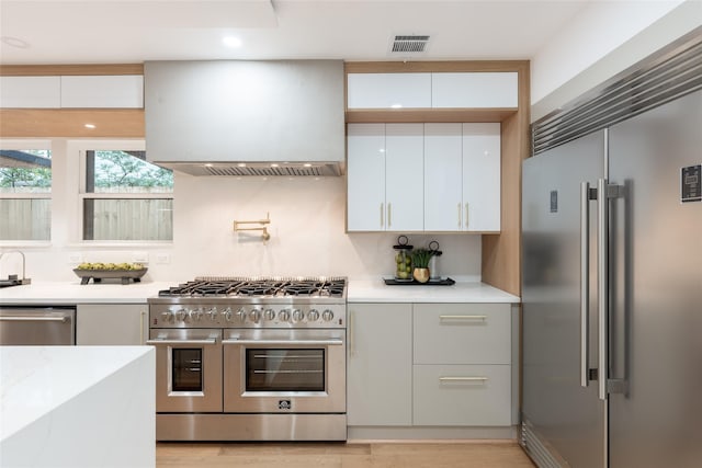 kitchen with white cabinets, high quality appliances, wall chimney range hood, and light hardwood / wood-style flooring