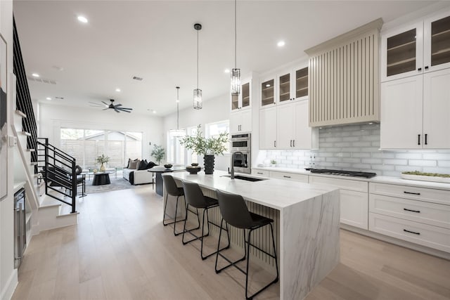 kitchen featuring light wood-type flooring, tasteful backsplash, a kitchen island with sink, ceiling fan, and sink