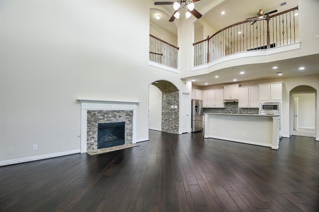 unfurnished living room with ceiling fan, sink, dark wood-type flooring, a stone fireplace, and a towering ceiling