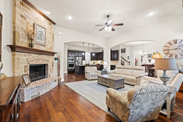 living room with ceiling fan with notable chandelier, a stone fireplace, and dark wood-type flooring