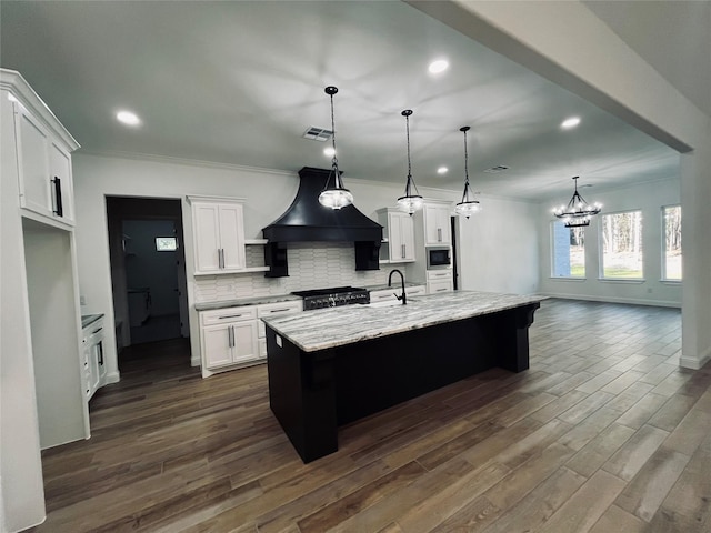 kitchen with stainless steel microwave, an island with sink, white cabinets, hanging light fixtures, and custom range hood