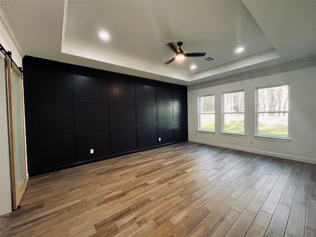 unfurnished bedroom featuring ceiling fan, a barn door, light hardwood / wood-style floors, and a tray ceiling