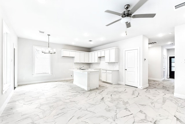 kitchen featuring decorative backsplash, ceiling fan with notable chandelier, white cabinetry, hanging light fixtures, and an island with sink