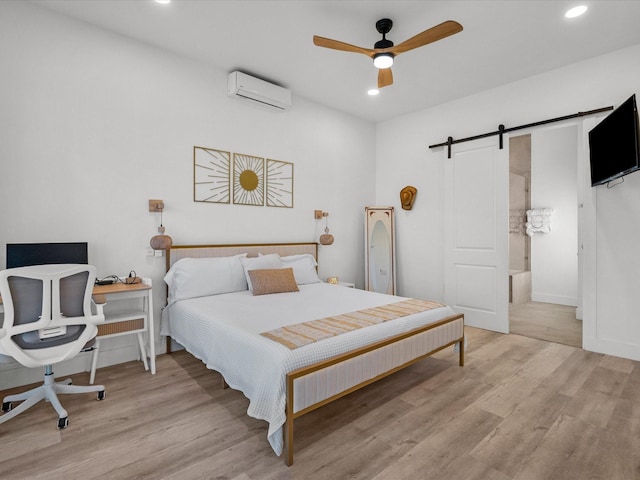 bedroom featuring ceiling fan, a barn door, light hardwood / wood-style flooring, an AC wall unit, and connected bathroom