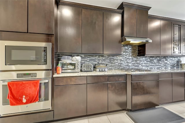 kitchen featuring decorative backsplash, light tile patterned floors, appliances with stainless steel finishes, dark brown cabinets, and light stone counters