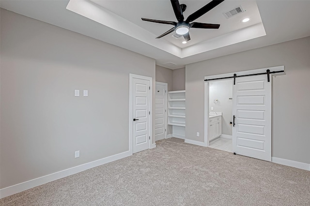 unfurnished bedroom featuring connected bathroom, ceiling fan, a barn door, light colored carpet, and a tray ceiling