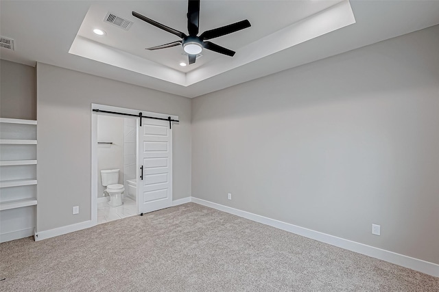 unfurnished bedroom with light carpet, a tray ceiling, ceiling fan, a barn door, and connected bathroom