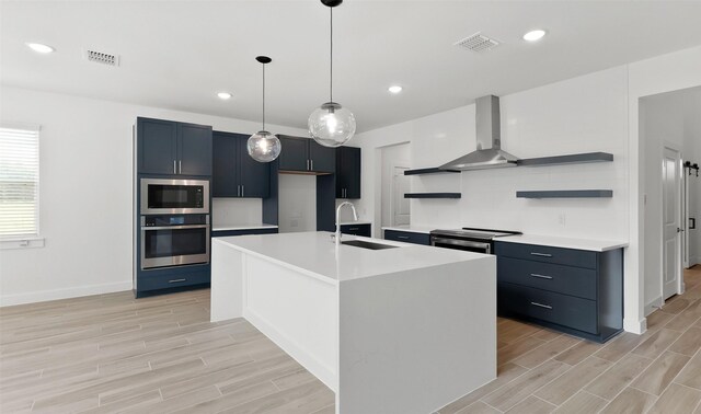 kitchen featuring sink, stainless steel appliances, wall chimney range hood, light hardwood / wood-style floors, and a kitchen island with sink