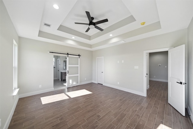 unfurnished bedroom with dark hardwood / wood-style flooring, a raised ceiling, ceiling fan, a barn door, and connected bathroom