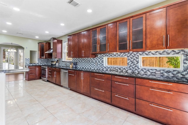 kitchen with decorative backsplash, wall chimney exhaust hood, stainless steel appliances, light tile patterned floors, and dark stone countertops