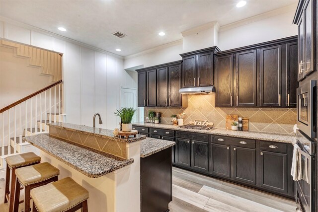 kitchen featuring stainless steel appliances, under cabinet range hood, a kitchen bar, and light stone countertops