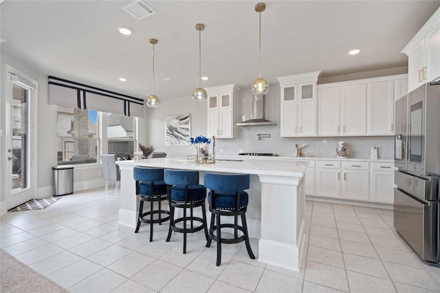 kitchen with a kitchen island with sink, white cabinets, hanging light fixtures, wall chimney exhaust hood, and stainless steel fridge