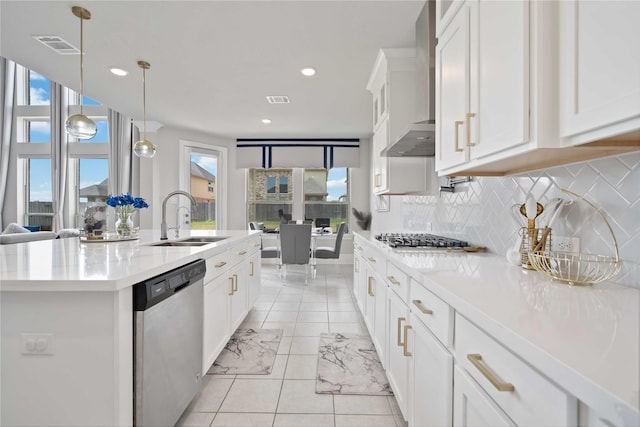 kitchen with sink, wall chimney exhaust hood, stainless steel appliances, decorative light fixtures, and a center island with sink