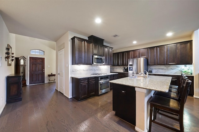 kitchen with light stone countertops, stainless steel appliances, a kitchen island with sink, dark hardwood / wood-style floors, and a breakfast bar area