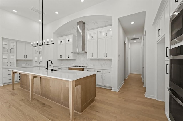 kitchen with sink, white cabinets, hanging light fixtures, a kitchen island with sink, and wall chimney exhaust hood