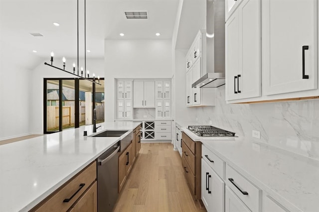 kitchen featuring wall chimney range hood, white cabinetry, hanging light fixtures, stainless steel appliances, and light stone counters