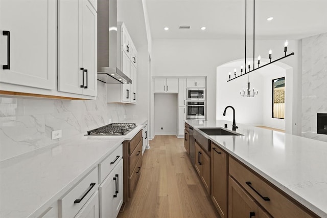 kitchen featuring wall chimney exhaust hood, light stone counters, hanging light fixtures, stainless steel appliances, and white cabinets