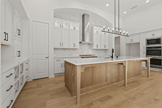 kitchen featuring wall chimney range hood, an island with sink, pendant lighting, stainless steel appliances, and white cabinets