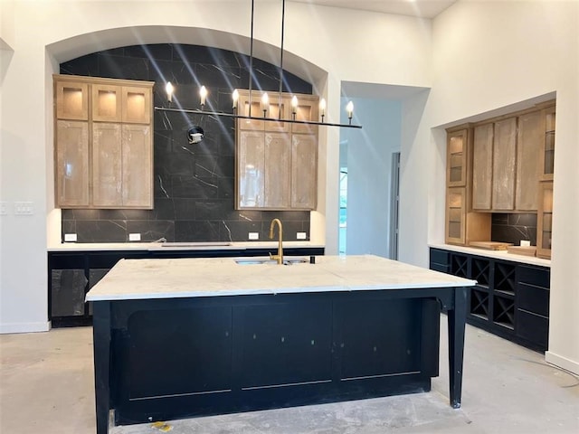 kitchen featuring an island with sink, decorative backsplash, light brown cabinetry, light stone countertops, and sink