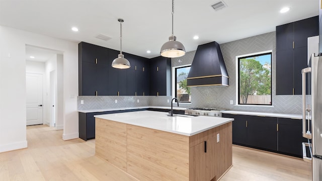 kitchen featuring light wood-type flooring, custom exhaust hood, sink, pendant lighting, and a center island with sink