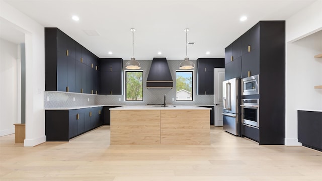 kitchen featuring custom exhaust hood, stainless steel appliances, pendant lighting, light hardwood / wood-style flooring, and an island with sink