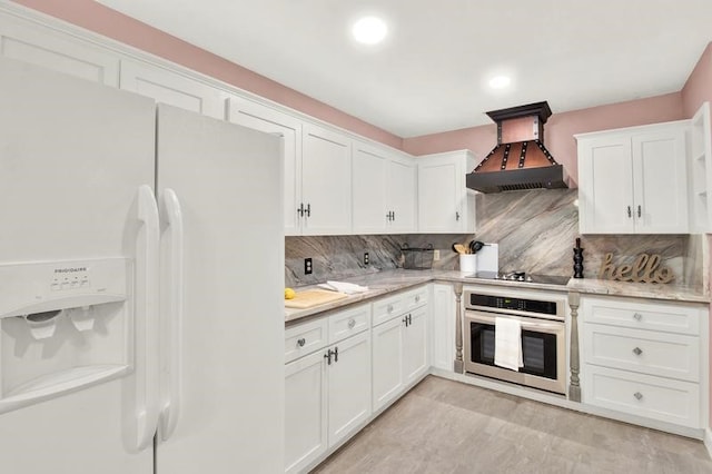 kitchen featuring custom exhaust hood, stainless steel oven, white refrigerator with ice dispenser, backsplash, and white cabinetry