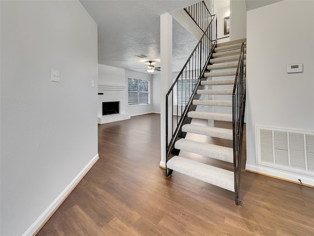 staircase featuring hardwood / wood-style flooring, ceiling fan, a textured ceiling, and a brick fireplace