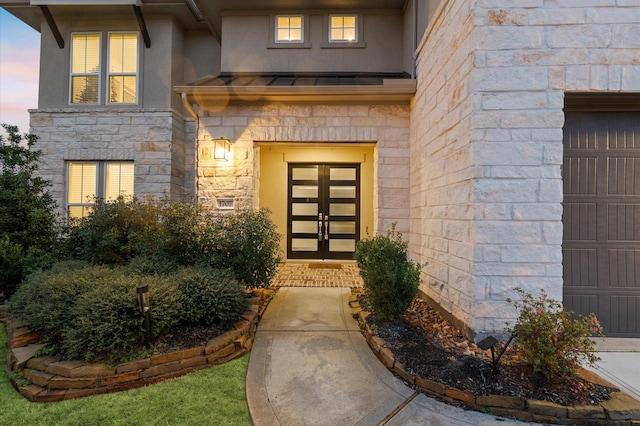 exterior entry at dusk featuring a garage, stone siding, french doors, stucco siding, and a standing seam roof