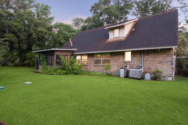 back of house at dusk featuring a sunroom, central AC unit, a lawn, and brick siding