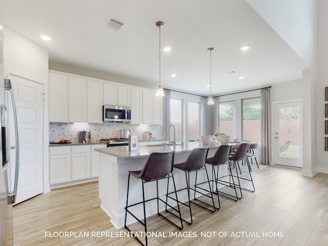 kitchen with white cabinets, hanging light fixtures, an island with sink, light hardwood / wood-style floors, and stainless steel appliances