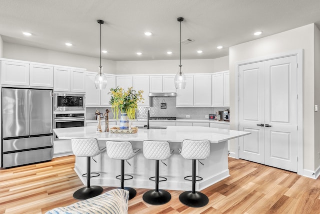 kitchen featuring a kitchen island with sink, light wood-type flooring, decorative light fixtures, white cabinetry, and stainless steel appliances