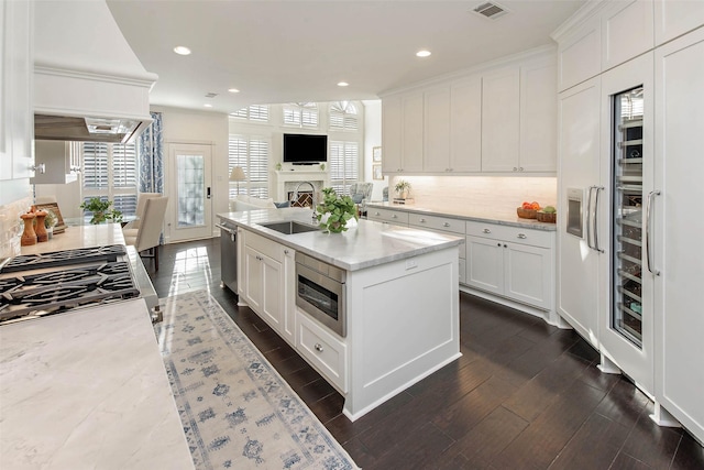 kitchen with premium range hood, sink, light stone countertops, an island with sink, and white cabinetry