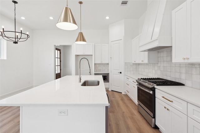 kitchen with sink, stainless steel gas range, premium range hood, pendant lighting, and a center island with sink