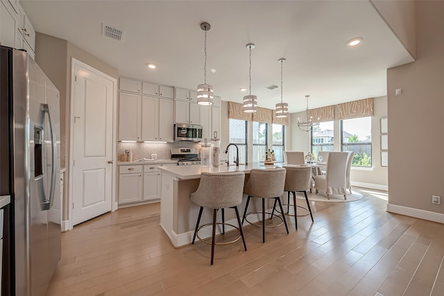 kitchen featuring light hardwood / wood-style flooring, an island with sink, appliances with stainless steel finishes, decorative light fixtures, and white cabinetry