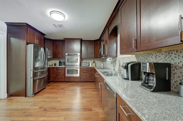 kitchen with appliances with stainless steel finishes, backsplash, light stone counters, sink, and light hardwood / wood-style flooring