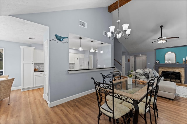 dining space featuring lofted ceiling with beams, a stone fireplace, ceiling fan with notable chandelier, and light hardwood / wood-style flooring